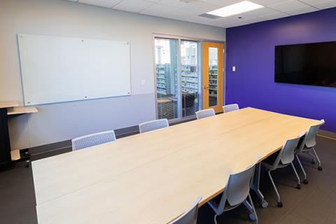 A conference room set up with tables and chairs with a tv screen and dry erase board on the walls.