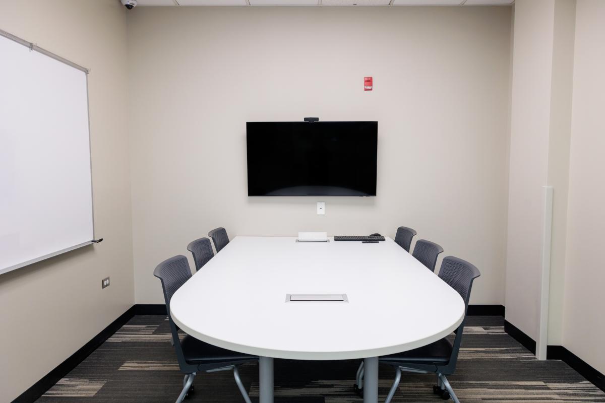 A conference room set up with tables and chairs with a tv screen and a white board on the walls..